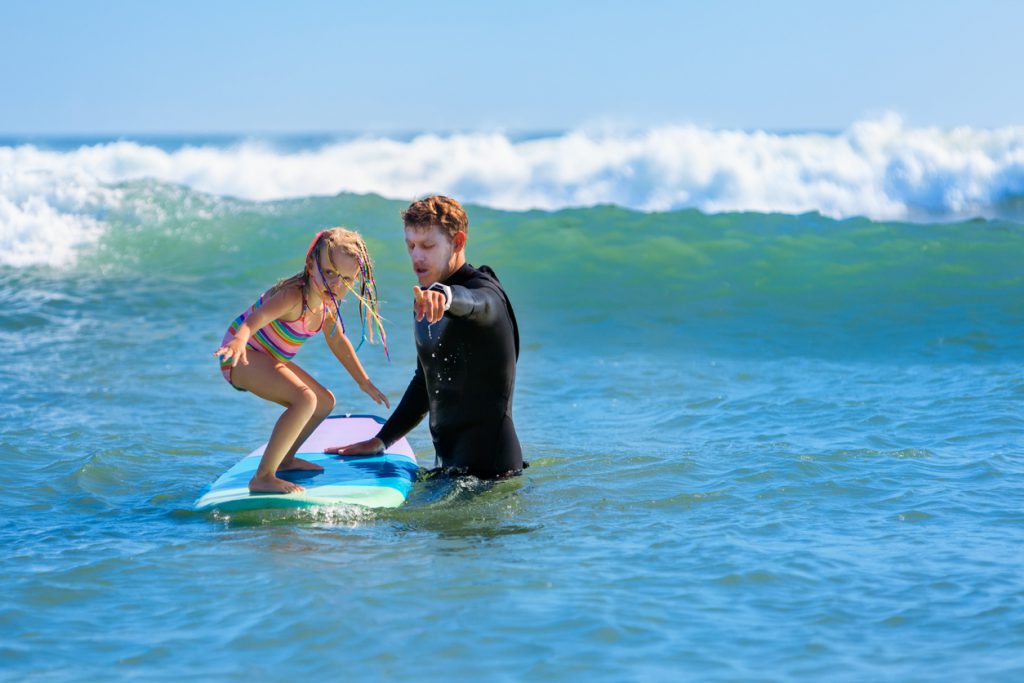 Little surf girl - a young surfer learns to ride on a surfboard with the instructor at surfing school. Active family lifestyle, kids water sports lessons, Vacation with child.